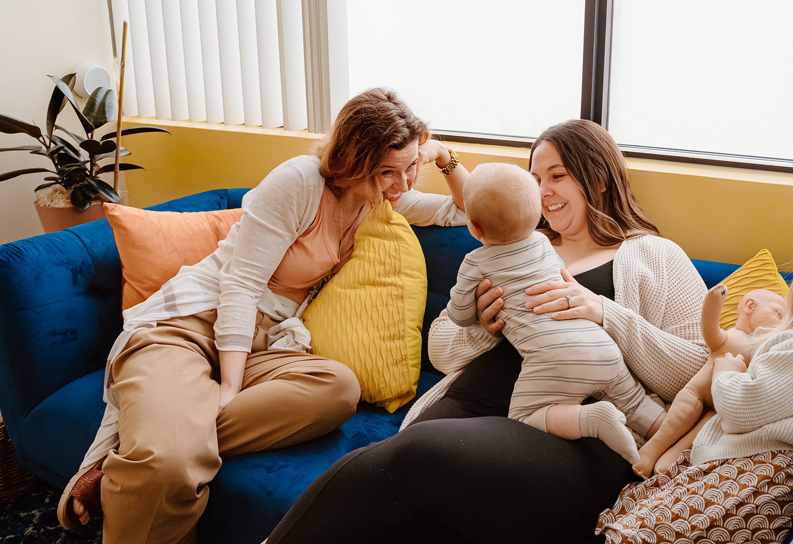 Rachel flirts with a baby in his mother's lap on couch by window